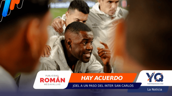 Soccer players in gray training suits huddle together, one man in front speaking with a raised finger, sponsor banners overlaid at the bottom.