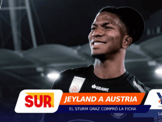 Smiling male athlete in a black sports jersey at a stadium, with colorful sponsor banners overlaying the lower part of the image (branding visible).