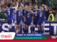 Japanese futsal team in blue jerseys celebrate on the court, high-fiving and smiling as fans cheer in the background