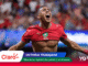 Moroccan footballer celebrating a goal with arms outstretched in a red jersey, stadium crowd in the background.