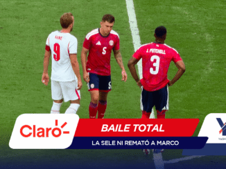 Three soccer players stand on a green pitch near a white line, two in red uniforms and one in white, with sponsor banners overlapping the lower edge of the image.
