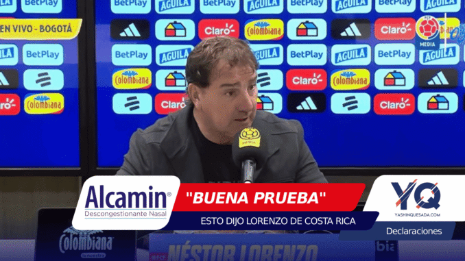 Man speaking at a press conference with a blue sponsor backdrop and a yellow mic flag in front of him, logos visible behind him (Colombiana, Claro, BetPlay).
