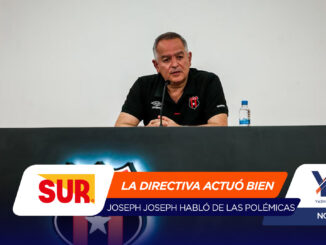 Man in a black polo seated at a press conference table with a microphone in front and a water bottle to the right, against a plain backdrop.