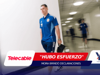 Male athlete in a blue training jersey walks down a hallway, carrying a small black bag in his right hand, with a second blurred figure in the background and bold sponsor banners across the bottom of the frame.