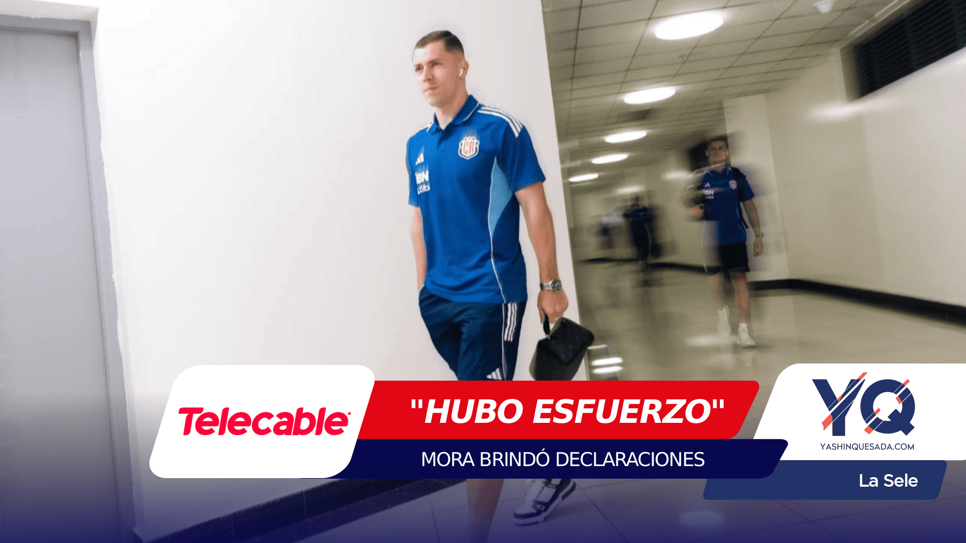 Male athlete in a blue training jersey walks down a hallway, carrying a small black bag in his right hand, with a second blurred figure in the background and bold sponsor banners across the bottom of the frame.