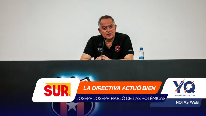 Man in a black polo seated at a press conference table with a microphone in front and a water bottle to the right, against a plain backdrop.