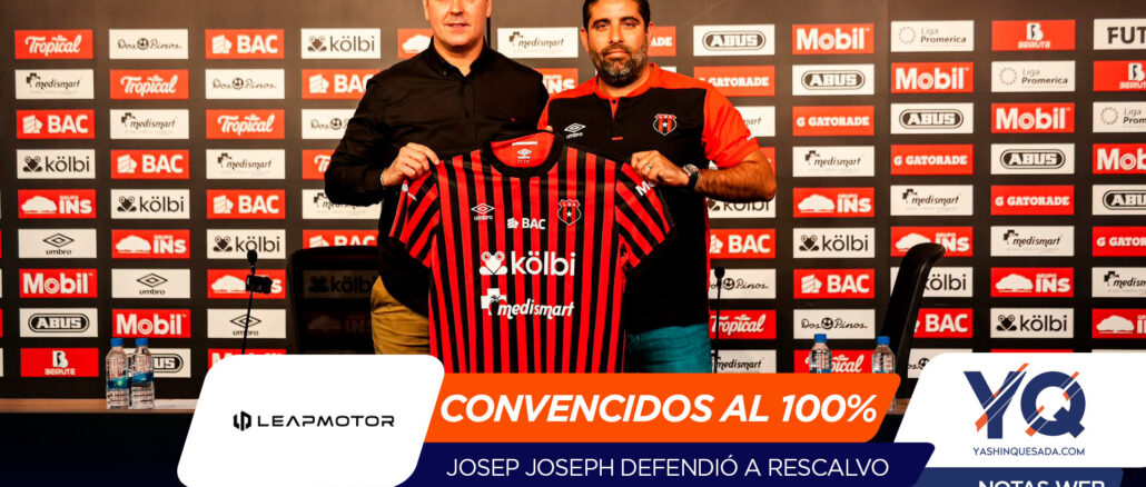 Two men at a press event hold up a red-and-black striped football jersey in front of a sponsor wall, presenting the club kit.