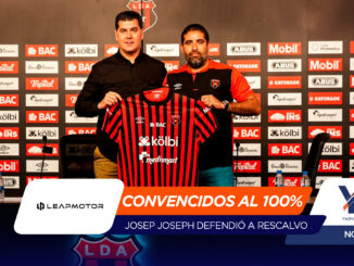 Two men at a press event hold up a red-and-black striped football jersey in front of a sponsor wall, presenting the club kit.