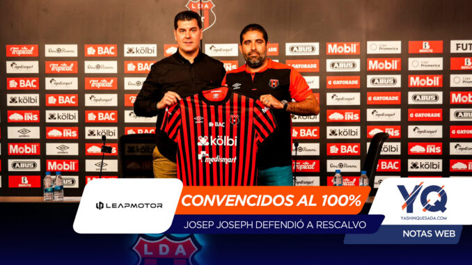 Two men at a press event hold up a red-and-black striped football jersey in front of a sponsor wall, presenting the club kit.