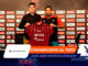 Two men at a press event hold up a red-and-black striped football jersey in front of a sponsor wall, presenting the club kit.