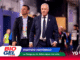 Two men in suits walking through a convention hall, with political banners and sponsor logos in the foreground (BIOGEL, Partido Histórico).