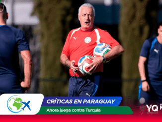 Coach in a red training shirt holds two soccer balls during a practice session, with players in blue shirts on a sunny field in the background.