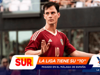 Soccer player in a maroon jersey with number 10 raises a hand on the field, with colorful sponsor banners across the bottom.