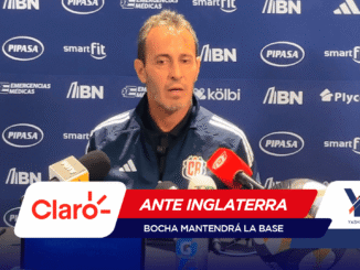 Man in a blue tracksuit speaking at a press conference with multiple microphones in front of a blue sponsor backdrop.