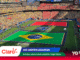 Bird's-eye view of a packed stadium with a large Brazilian flag on the field and teams lined up for a ceremony amidst colorful stands and banners.