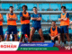 Group of seven soccer players in blue kits with orange training bibs standing with arms crossed on a stadium pitch; promo banner at bottom.
