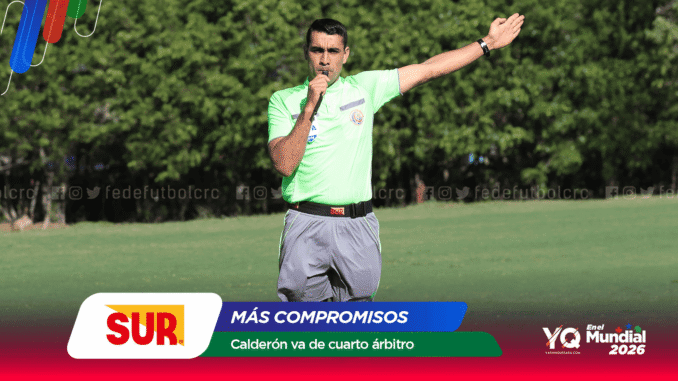 Male soccer referee in a neon green shirt blows a whistle and points with his right arm extended on a grassy field, with sponsor banners in the foreground.