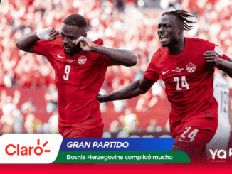 Two Canadian soccer players in red celebrate a goal on the field as a cheering crowd looks on, with sponsor banners at the bottom of the image.