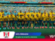 Ecuador national soccer team wearing yellow jerseys and blue shorts posing for a group photo on the pitch with a full stadium in the background.