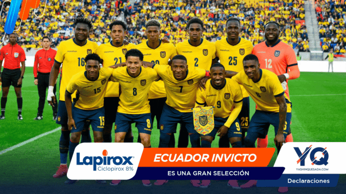 A team of Ecuadorian soccer players in yellow jerseys poses for a group photo on a green field with a cheering crowd behind them, including a pink-clad goalkeeper on the right.