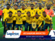 A team of Ecuadorian soccer players in yellow jerseys poses for a group photo on a green field with a cheering crowd behind them, including a pink-clad goalkeeper on the right.