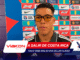 Man wearing black-rimmed glasses being interviewed at a press conference in front of a colorful sponsor backdrop.