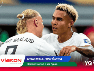 Two footballers in white jerseys embrace in a celebratory moment on the field, with sponsor banners and text overlays in the foreground.