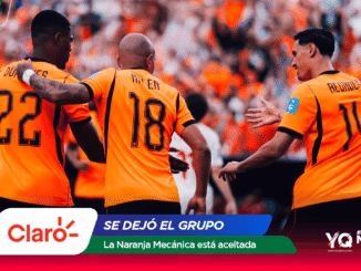 Three soccer players in orange jerseys celebrate together on the field as a cheering crowd looks on.