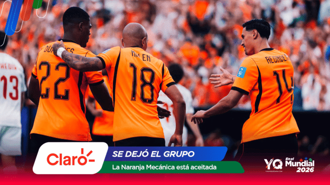 Three soccer players in orange jerseys celebrate together on the field as a cheering crowd looks on.