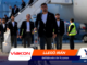 Men in suits walking through an airport tarmac, leading luggage carts; security staff in neon vests in background.