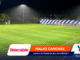 Soccer field at night illuminated by floodlights with advertising banners along the edge of the pitch.