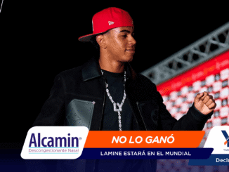 Male performer in a red cap on a dark stage, with sponsor banners reading 'Alcamín' and 'NO LO GANÓ' in the foreground.