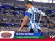 Soccer player in a blue-and-white striped jersey on a packed stadium field with Roma and Mundial 2026 banners in view.