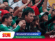 Energetic crowd of sports fans wearing green, cheering and raising their arms at a stadium; SUR logo and Ecuador banners visible at the bottom.