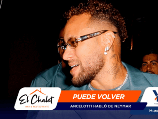 Close-up of a man with curly hair and beard, wearing sunglasses and earrings, at a dark event with orange-blue promotional banners reading 'PUEDE VOLVER' in the foreground.