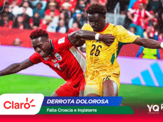 Two football players in red and yellow kits jostle for the ball during a crowded match, with colorful sponsor banners along the bottom.