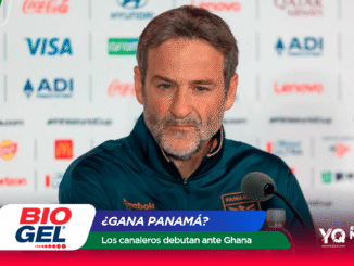 Man at a press conference speaking into a microphone, wearing a dark teal sports jacket, with sponsor logos on the backdrop and foreground banner obscuring part of the view.