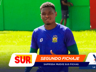 Portrait of a smiling male soccer player in a blue jersey on a sunny grassy field, with vibrant sponsor banners overlaid (SUR, SEGUNDO FICHAJE).