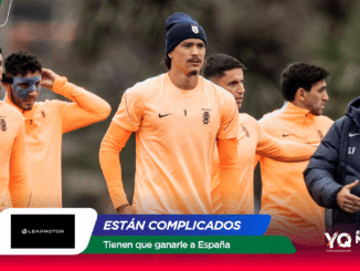 Equipo de fútbol entrenando al aire libre con camisetas naranja; varios jugadores de pie y un entrenador a la derecha con chaqueta oscura.