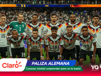 German national football team posing for a group photo on the pitch with fans in the stands behind them, players in white jerseys with black, red, and gold accents.