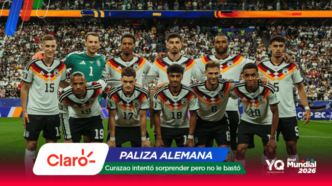 German national football team posing for a group photo on the pitch with fans in the stands behind them, players in white jerseys with black, red, and gold accents.