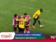 Group of soccer players in yellow and purple jerseys embrace in a huddle on a green pitch after a goal.
