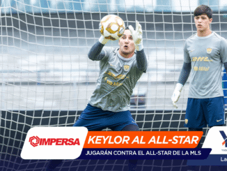 Two goalkeepers defend a soccer goal; one dives to catch a yellow ball while sponsor banners cover the bottom of the image.