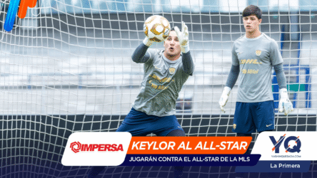 Two goalkeepers defend a soccer goal; one dives to catch a yellow ball while sponsor banners cover the bottom of the image.