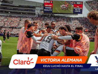 Soccer players in white and black celebrate a goal in a huddle on the field, with a crowded stadium and scoreboard in the background.