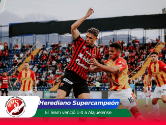 Soccer players duel for the ball on a crowded stadium pitch, with a banner announcing Herediano Supercampeón and the 1-0 victory text overlay.