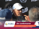 Man wearing a white cap speaks to reporters at a press conference with sponsor logos in the background.