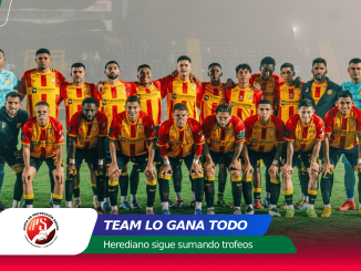 Group photo of a soccer team in red-and-yellow kits posing on a field, players standing and kneeling together with a stadium backdrop in the background.