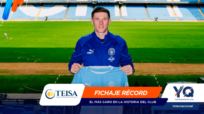 Man in a blue jacket holds up a light blue football jersey on a stadium pitch during a press event.