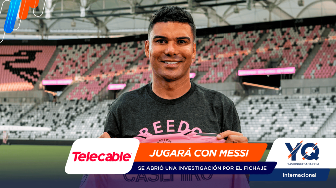 Man in a dark t-shirt at a football stadium, smiling and holding a pink shirt, with sponsor banners across the bottom.